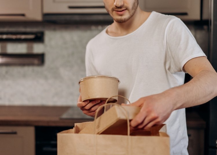 Man placing takeout containers into paper bag in kitchen setting.