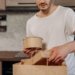 Man placing takeout containers into paper bag in kitchen setting.