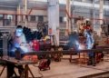 Two welders wearing protective gear working in a busy industrial factory workshop.