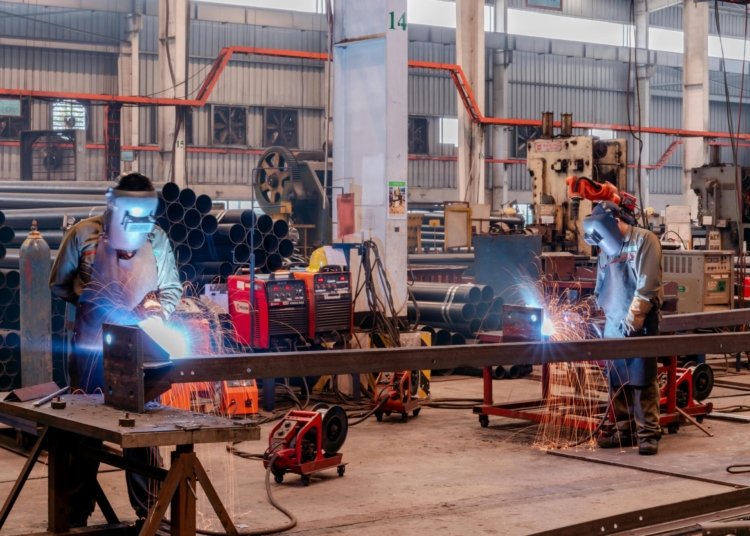 Two welders wearing protective gear working in a busy industrial factory workshop.