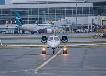 A private jet on the runway at a bustling airport with commercial planes and terminal in view.