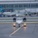 A private jet on the runway at a bustling airport with commercial planes and terminal in view.