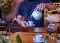 Barista expertly pouring hot tea into a white cup in a bustling cafe.