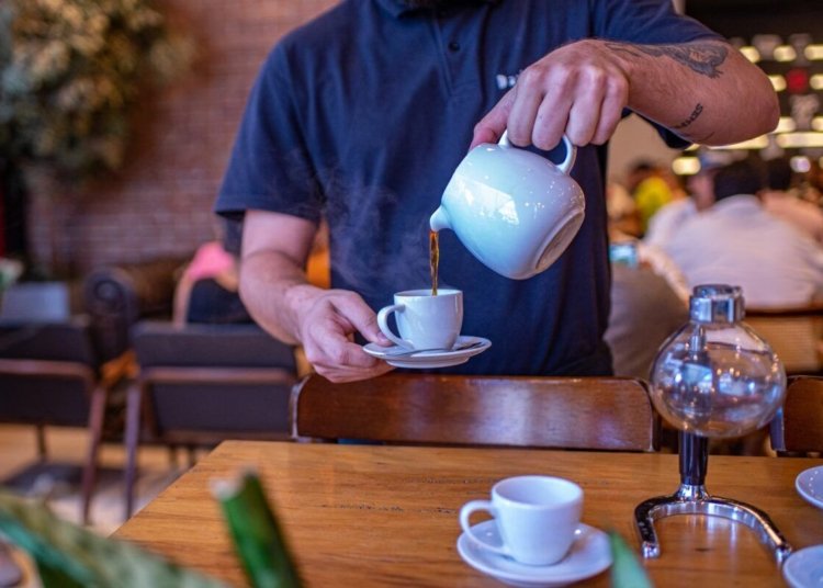 Barista expertly pouring hot tea into a white cup in a bustling cafe.