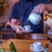 Barista expertly pouring hot tea into a white cup in a bustling cafe.