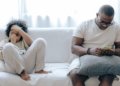 Concentrated African American man in nightwear using smartphone while sitting on cozy sofa with son resting with joystick in light room