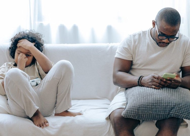 Concentrated African American man in nightwear using smartphone while sitting on cozy sofa with son resting with joystick in light room