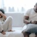 Concentrated African American man in nightwear using smartphone while sitting on cozy sofa with son resting with joystick in light room