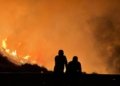 Two silhouettes observe a massive forest fire with intense flames and smoke filling the night sky.