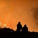 Two silhouettes observe a massive forest fire with intense flames and smoke filling the night sky.