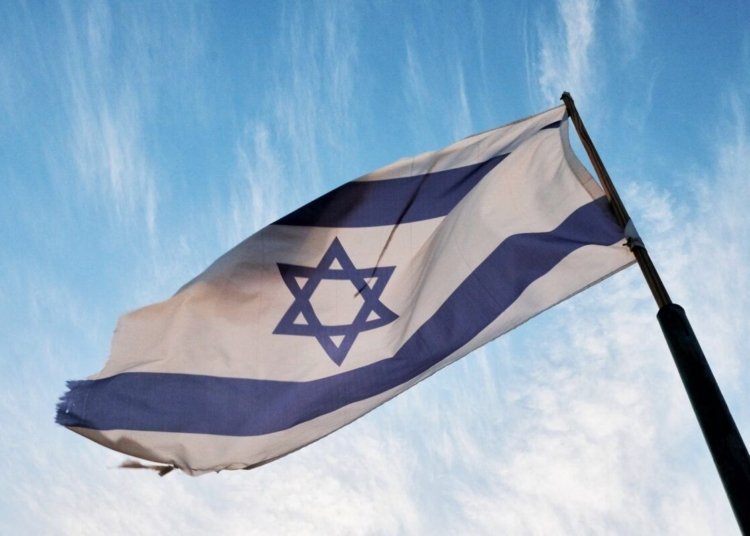 A close-up of the Israeli flag waving on a flagpole against a clear blue sky.