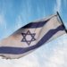 A close-up of the Israeli flag waving on a flagpole against a clear blue sky.