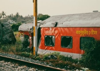 An abandoned and damaged train car rests beside tracks in a lush rural area.