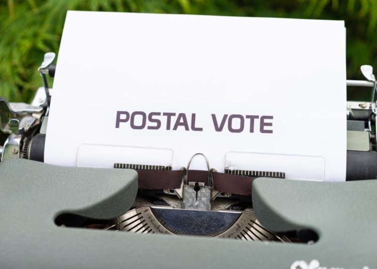 Close-up of typewriter with paper displaying the words 'Postal Vote', outdoors.